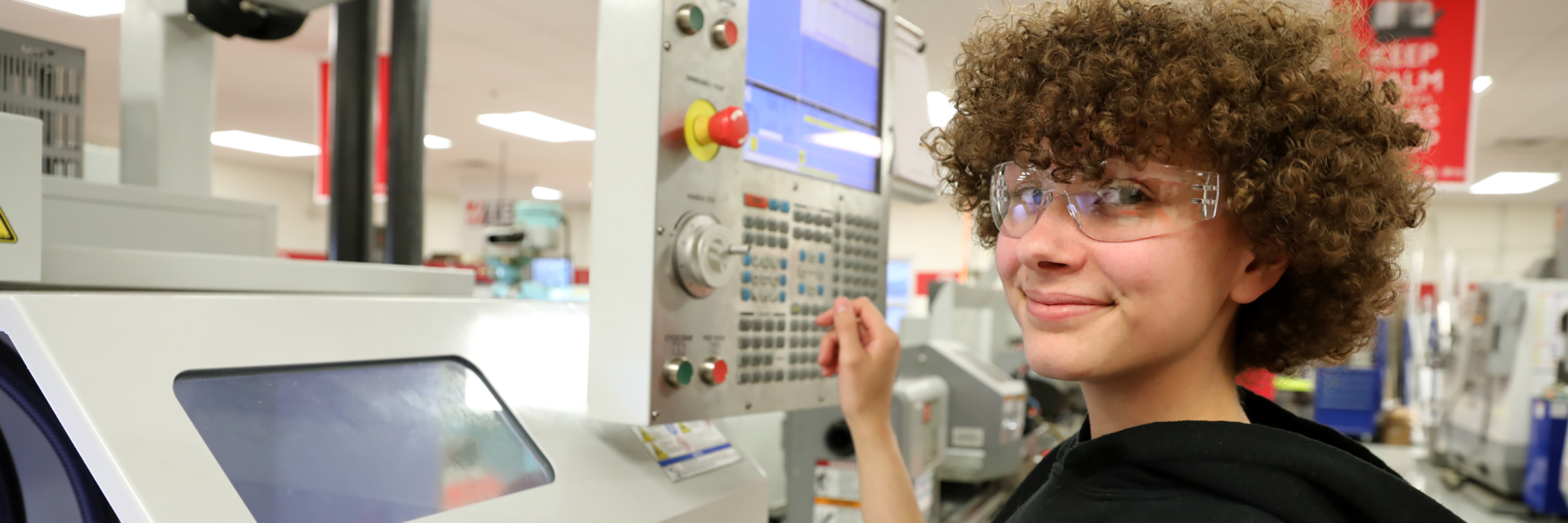 young person smiling at camera with safety goggles in a room full of machines